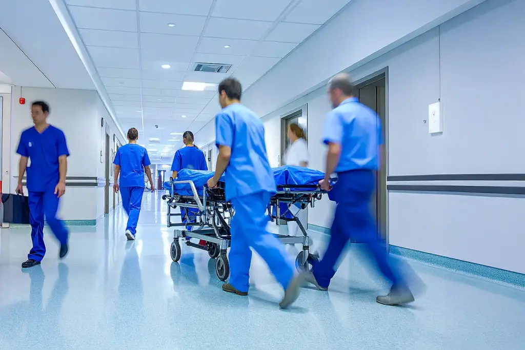 Busy hospital corridor with blurred healthcare staff and stretcher covered in blue, bright clinical lighting; urgent, dynamic medical scene.