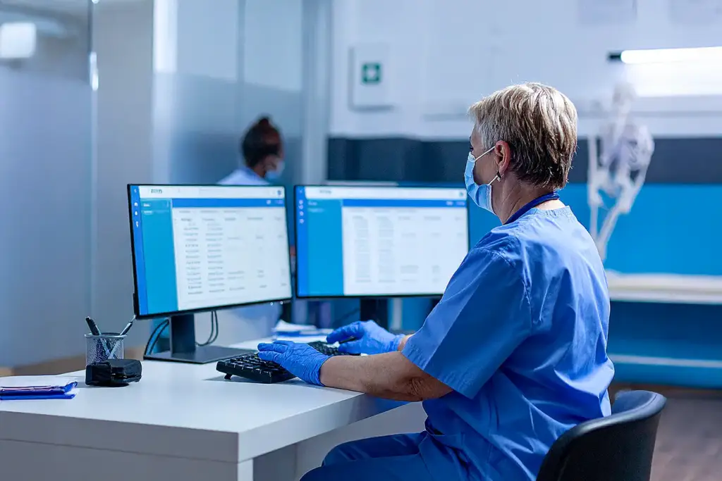 Female medical professional in blue uniform and gloves typing at workstation; blue-toned lab with monitors and medical equipment in background.